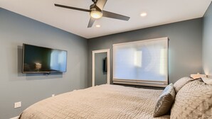 Une chambre à coucher avec un lit, un ventilateur de plafond, un téléviseur fixé au mur et une fenêtre avec des stores.