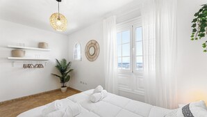 Ein modernes Schlafzimmer mit einem großen Bett, weißen Wänden, einer Zimmerpflanze und Blick durch ein Fenster.