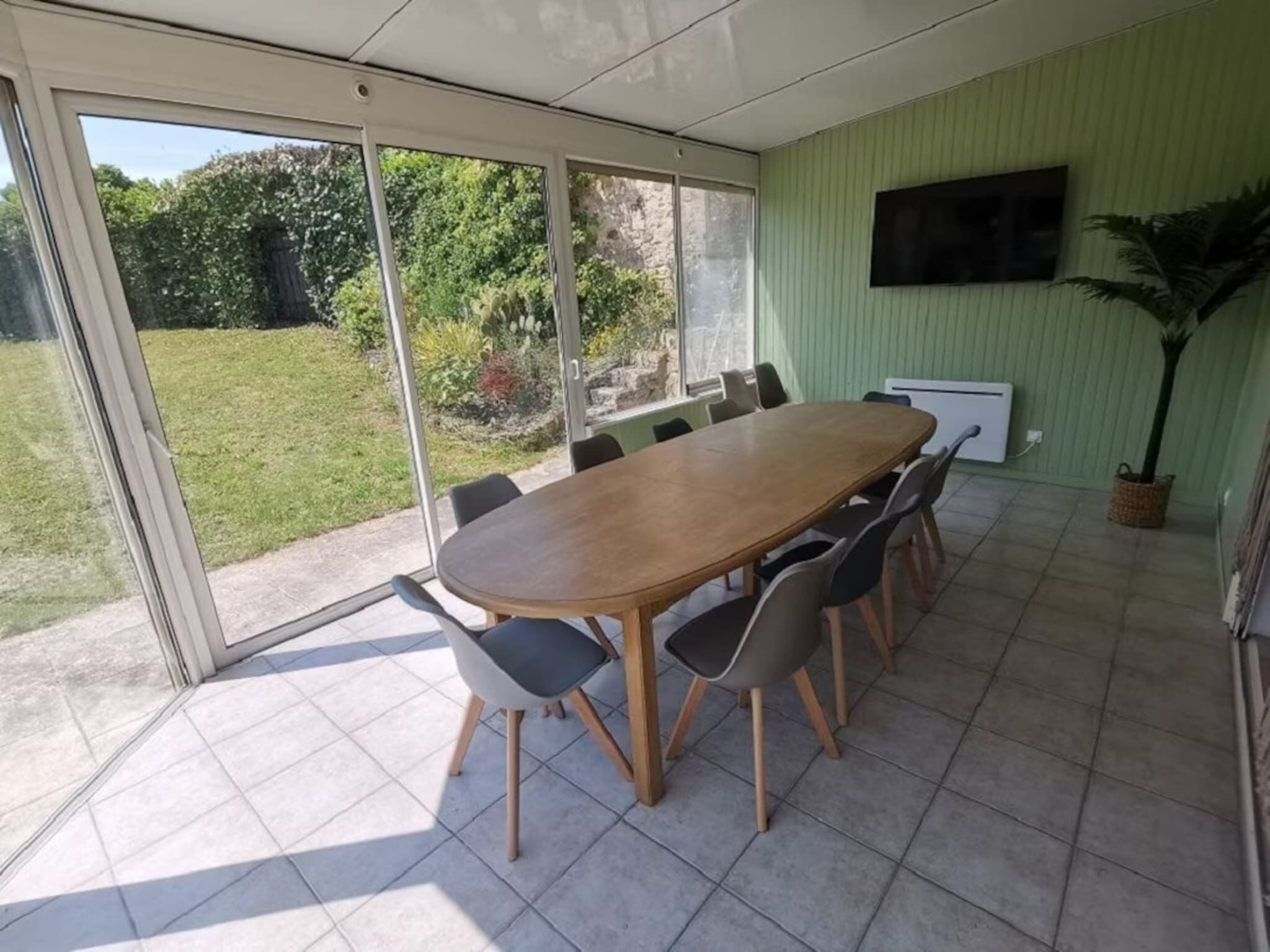 A dining area with a large table and chairs, a television mounted on the wall, and a view of the outdoors.