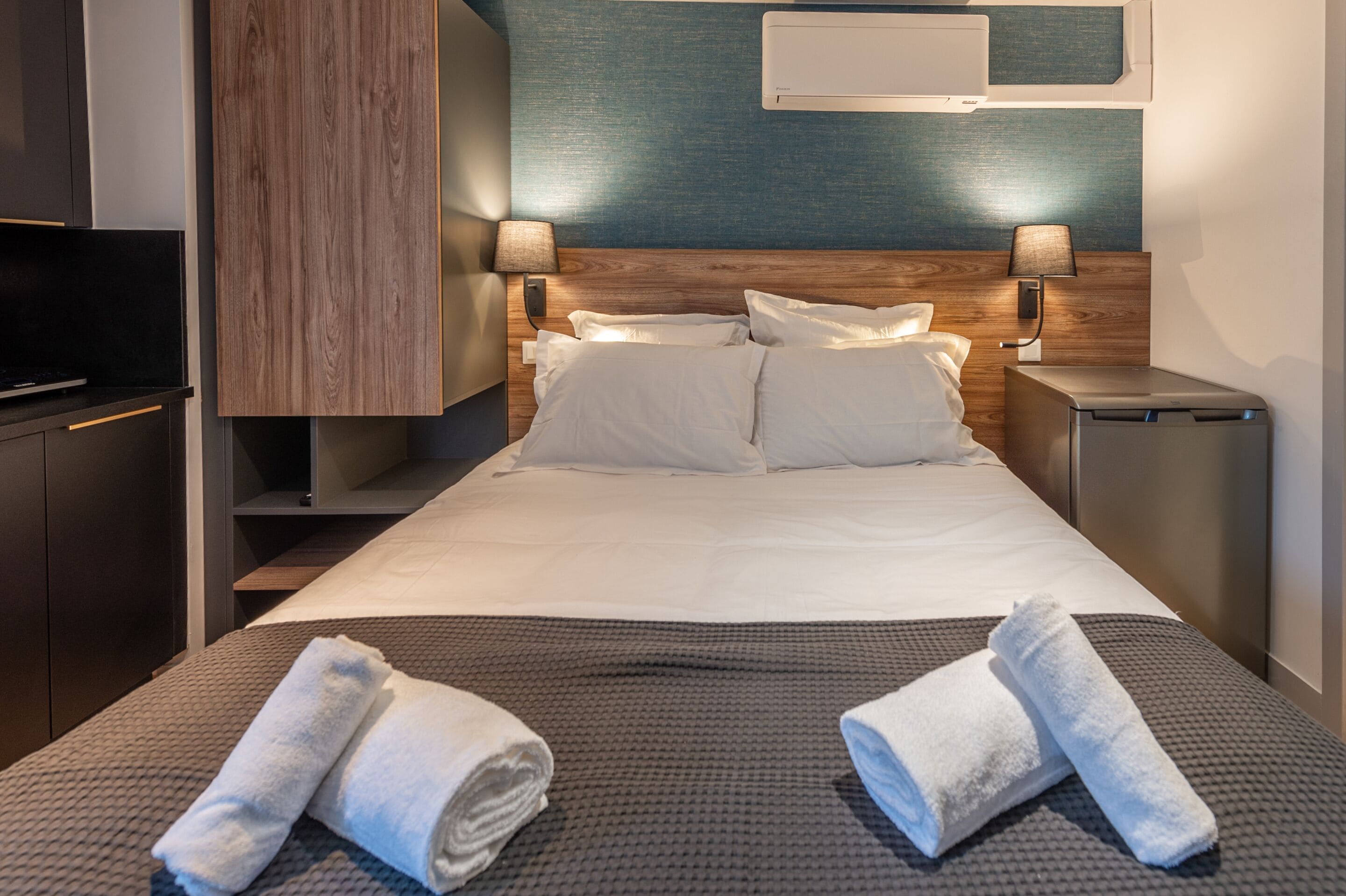 A modern hotel room with a neatly made bed, a flat-screen TV, a wooden headboard, and a wall-mounted air conditioning unit.