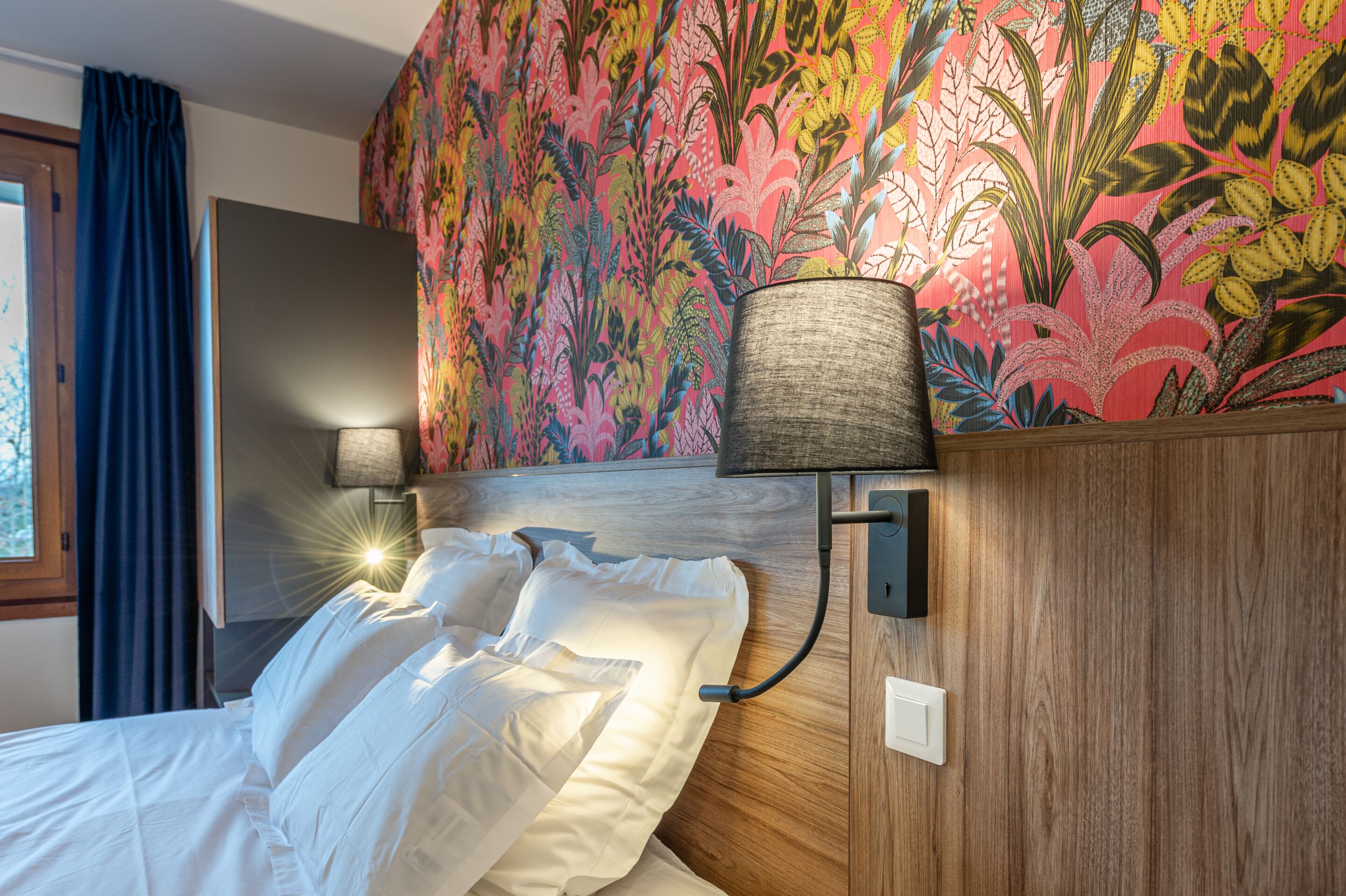 A hotel room with a bed, two bedside lamps, a floral wallpaper, and a window with curtains.
