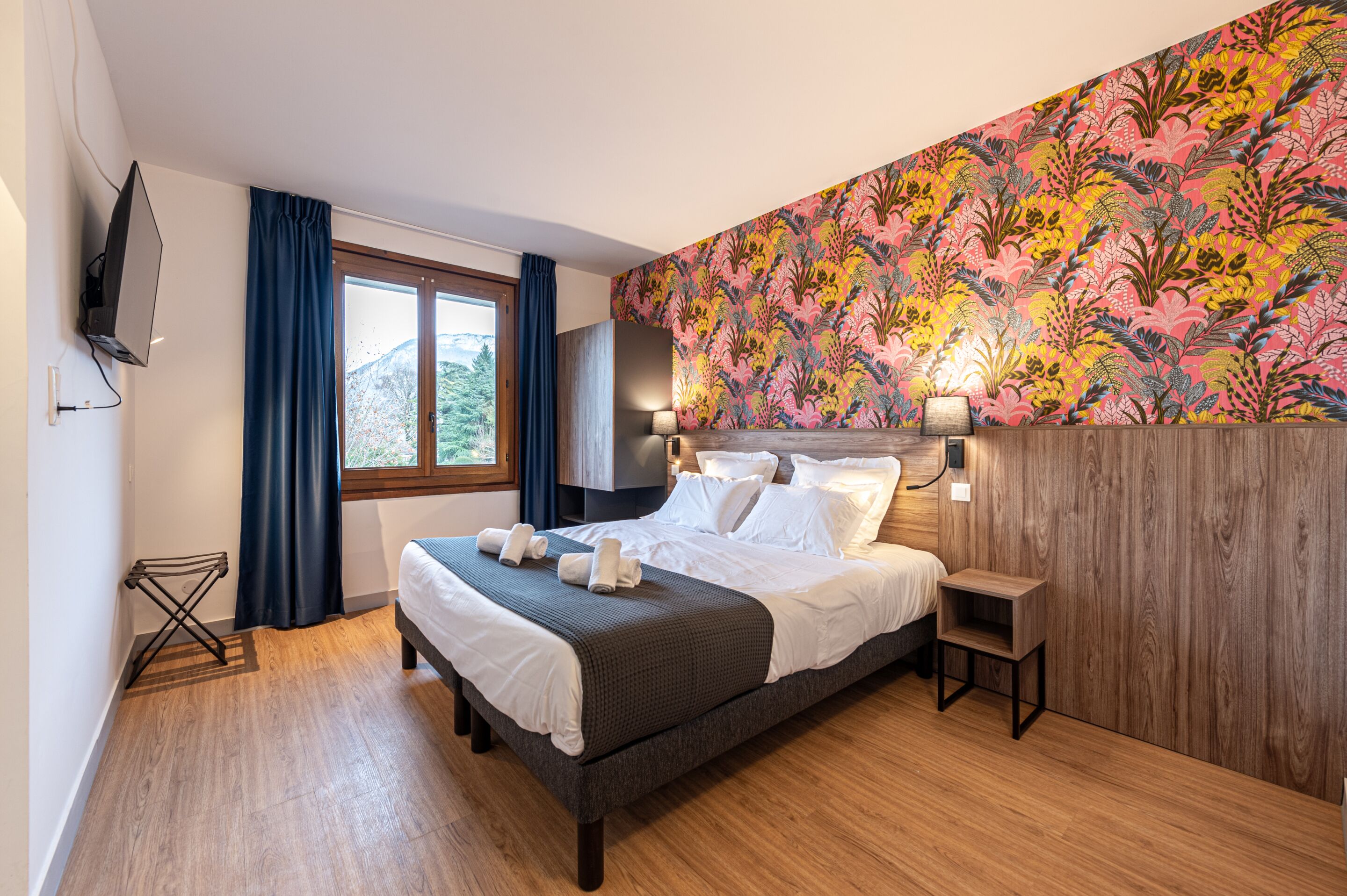 A modern hotel room with a large bed, a wooden headboard, a wall-mounted TV, and a window with curtains.