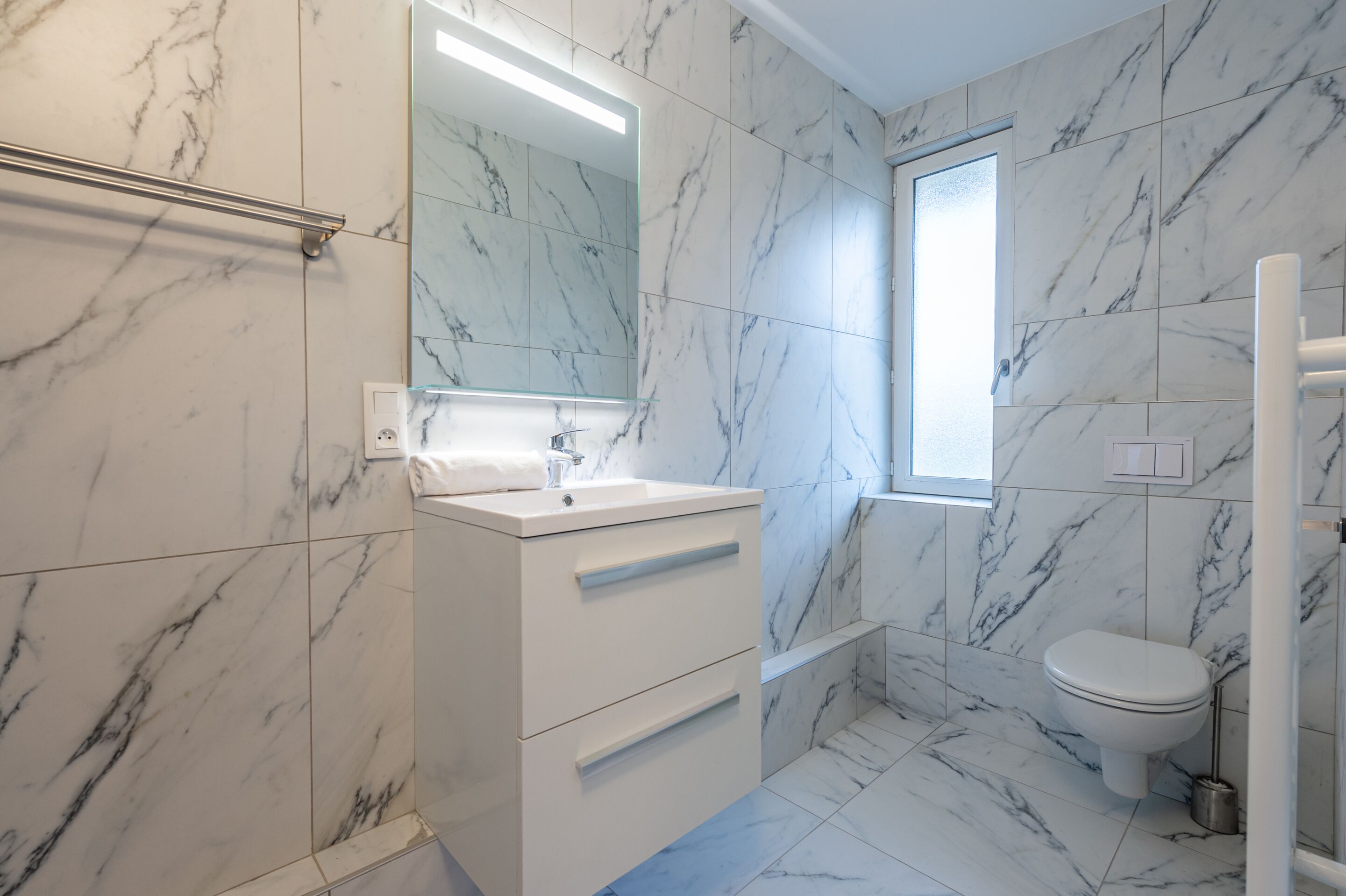Un baño moderno con baldosas de mármol, un tocador blanco con cajones, un espejo y un inodoro.