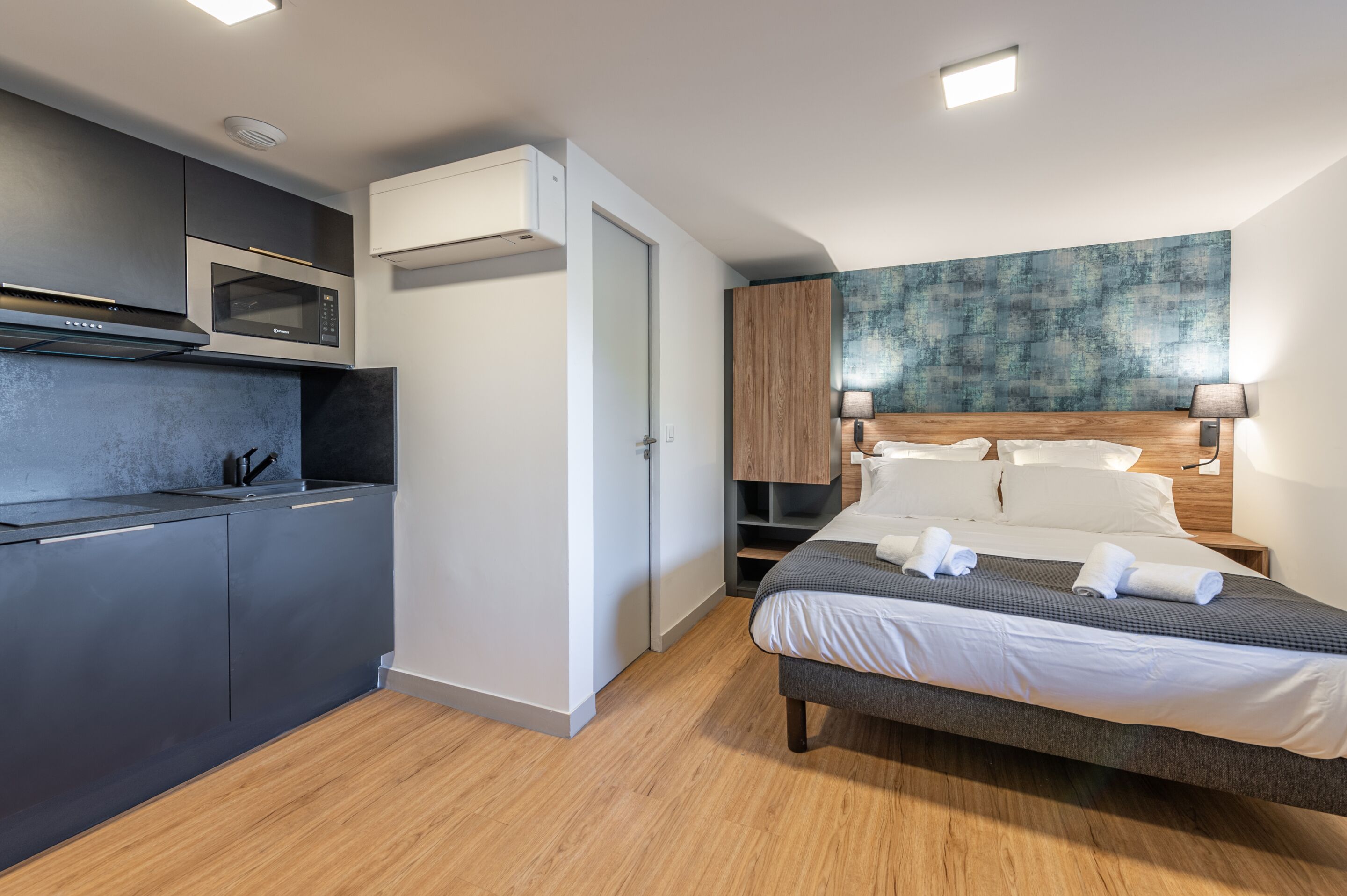 Ett modernt hotellrum med ett litet kök, en stor säng med prydligt vikta handdukar och ett väggkonstverk med en intressant struktur.