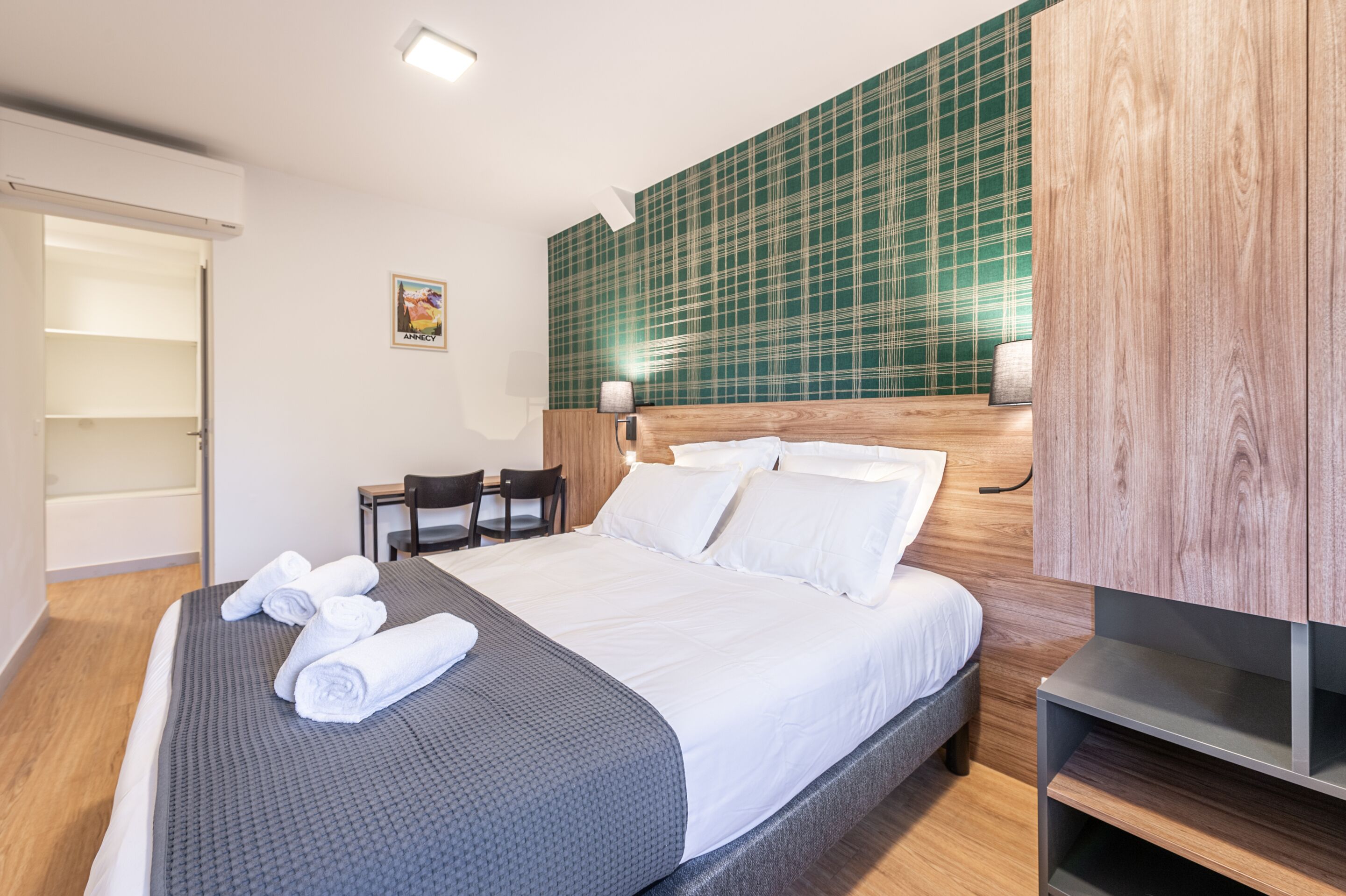 Ein Hotelzimmer mit einem großen Bett, einem hölzernen Kopfteil, einer Wand mit grün-kariertem Tapetenmuster, einem Schreibtisch mit Stuhl und einer Wandleuchte.