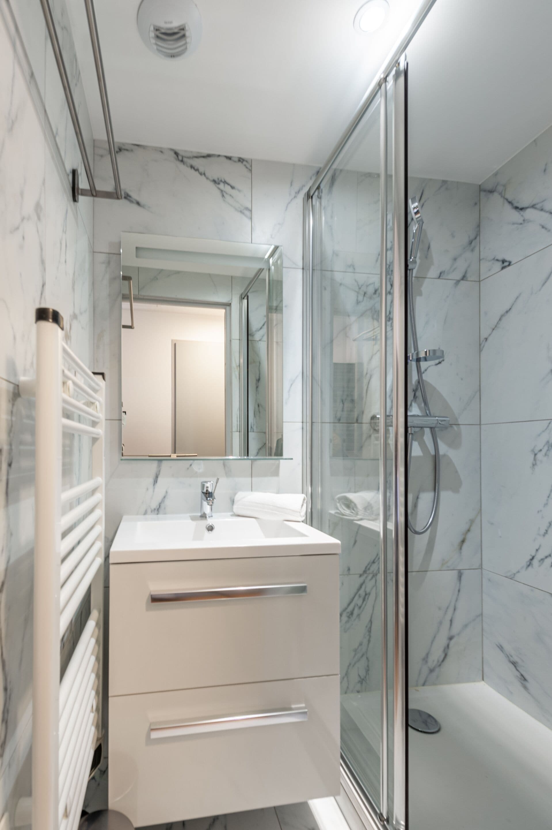 Een moderne badkamer met een douchecabine van glas, een wit wastafelmeubel met lades en een wand betegeld met marmer.