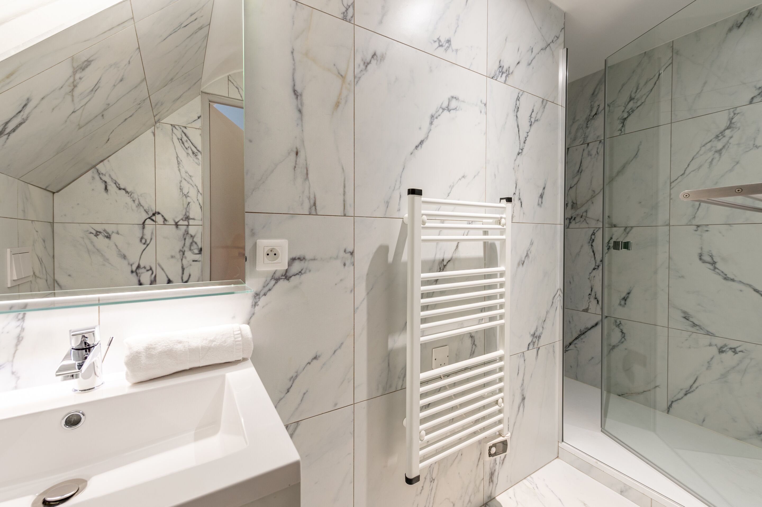 Une salle de bain moderne avec un mur recouvert de carreaux de marbre, un lavabo blanc et une douche séparée par une paroi de verre.