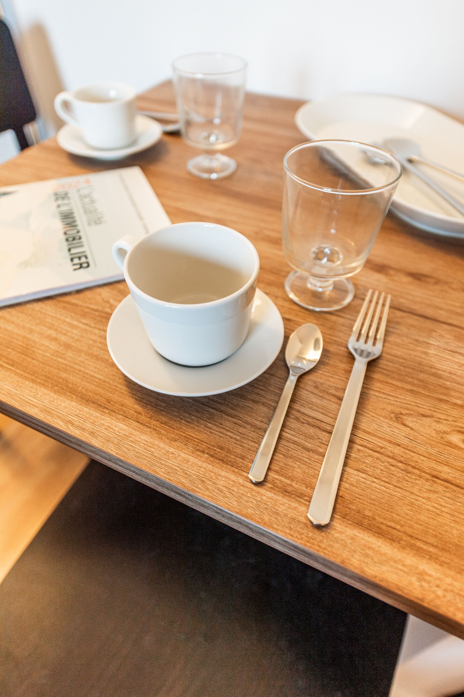 Een net gedekte eettafel met een witte kop, een glas, een lepel en een vork op een houten tafel.