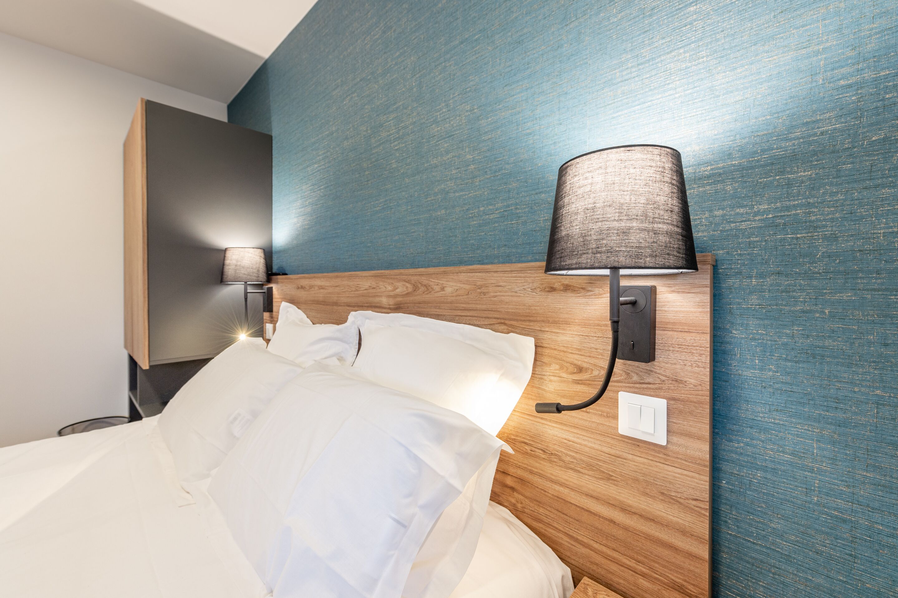 A neatly made bed with white linens and a wooden headboard. There is a wall-mounted lamp on the headboard and a small table beside the bed.