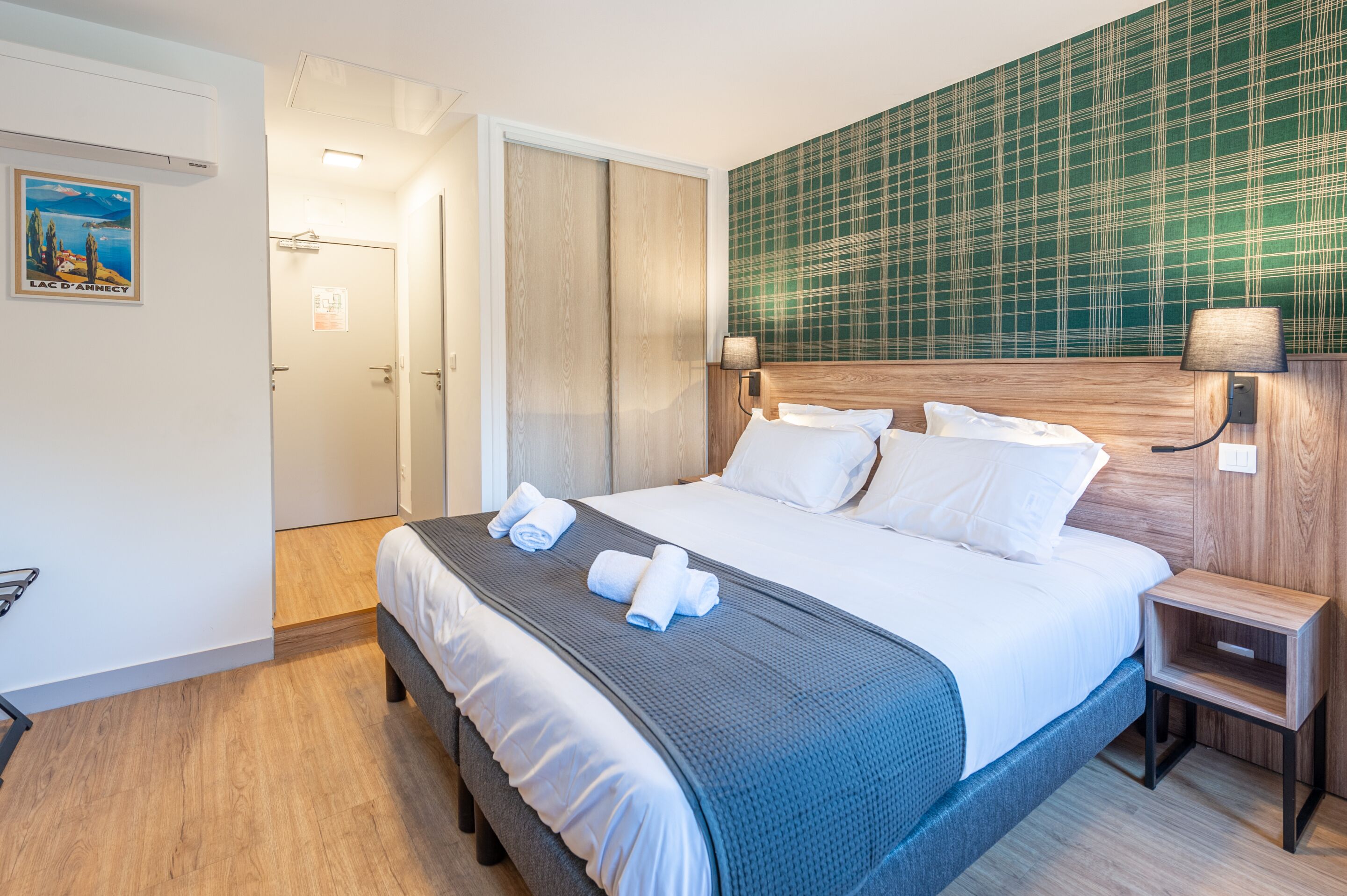 A hotel room with a large bed, a wooden headboard, a nightstand with a lamp, and a green plaid wallpaper.
