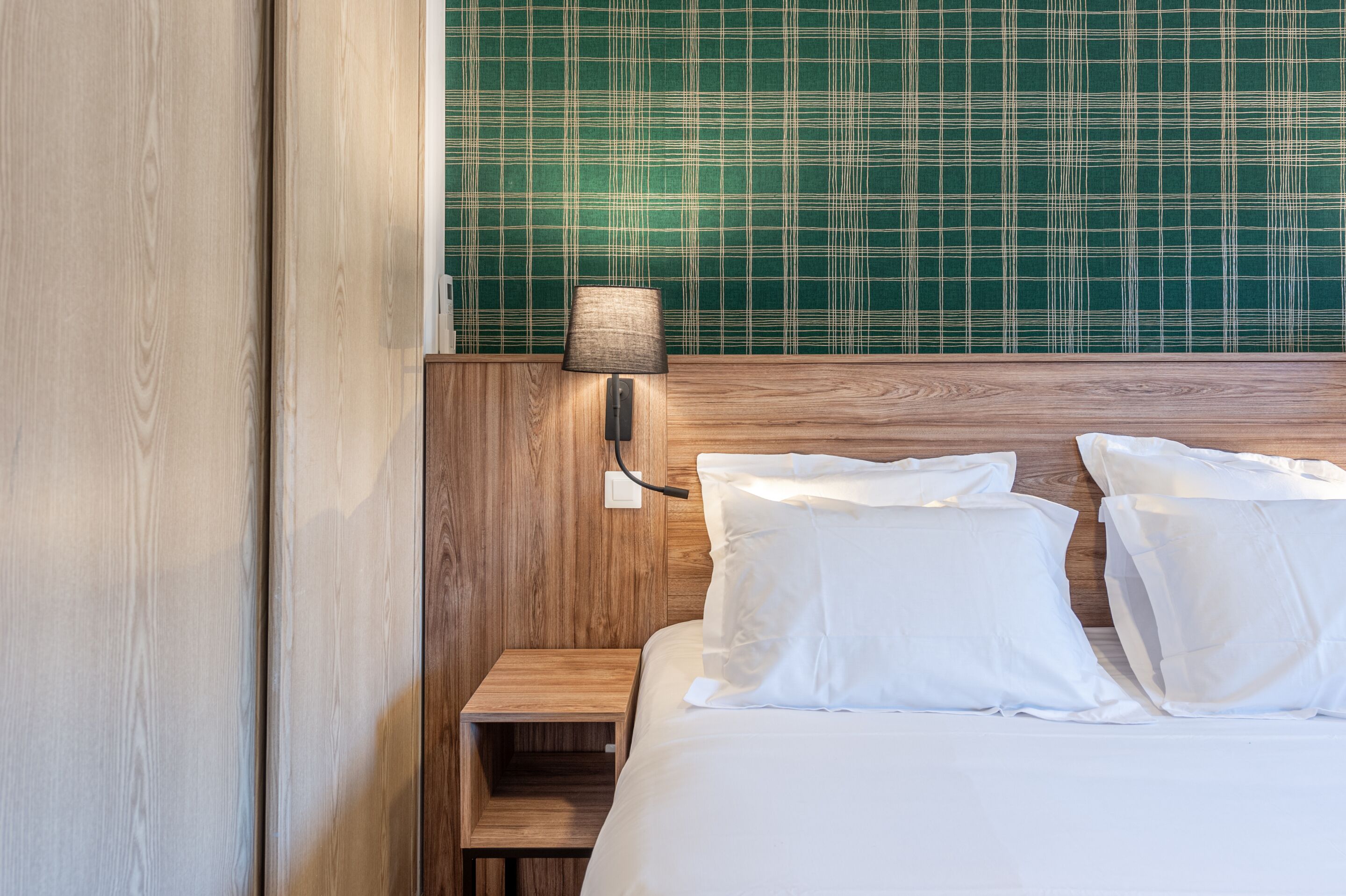 A neatly made bed with white linens and pillows, a wooden nightstand with a lamp, and a green plaid headboard.