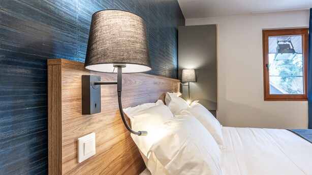 A hotel room with a wooden headboard, a wall-mounted lamp, a bedside table, and a window with a view of greenery. - Les Tonnelles (Sevrier)