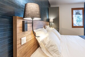 A hotel room with a wooden headboard, a wall-mounted lamp, a bedside table, and a window with a view of greenery. - Les Tonnelles (Sevrier)