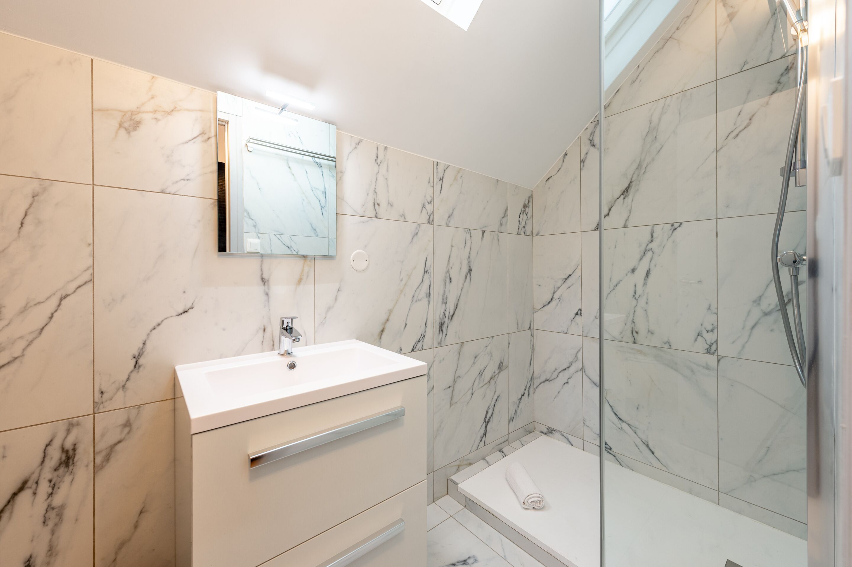 A bathroom with marble tiles, a white sink, a mirror, and a shower area.