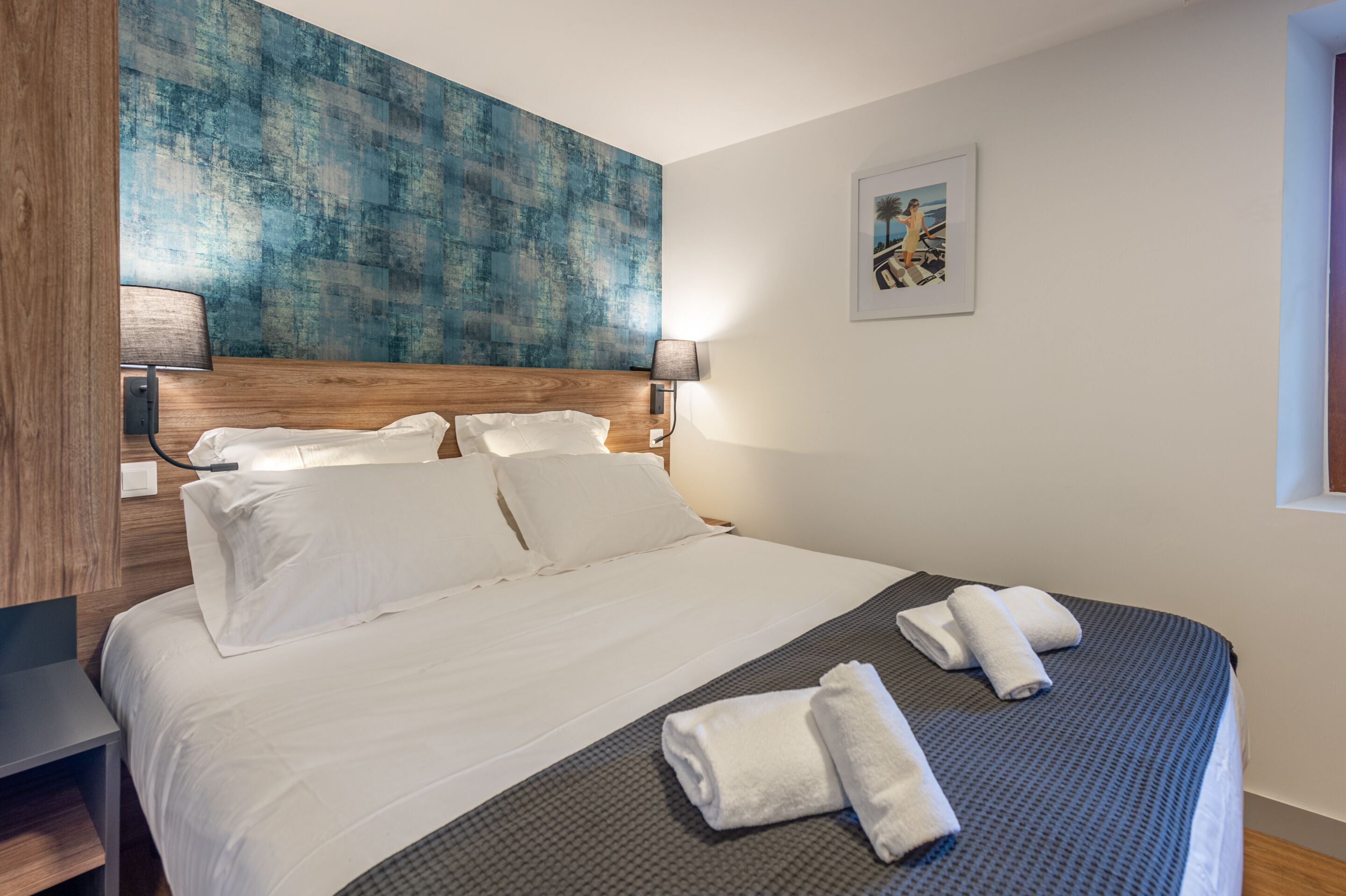 A hotel room with a large bed, white linens, two bedside lamps, a wooden headboard, a blue textured wall, and a framed picture on the wall.