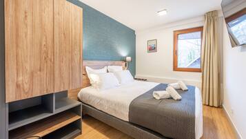 A modern hotel room with a bed, a wooden wardrobe, a blue accent wall, a window with curtains, and a framed picture on the wall.