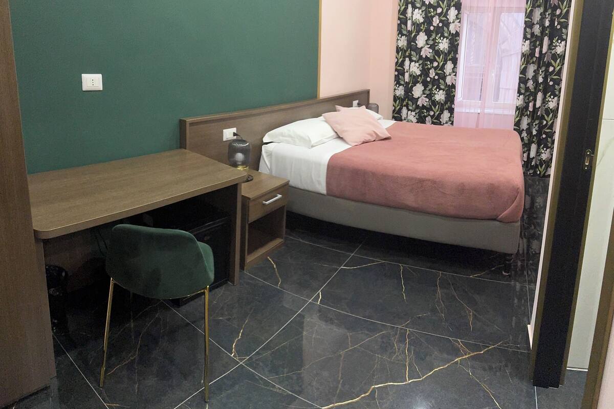 A hotel room with a bed, a desk, a chair, a nightstand, and a mirror.