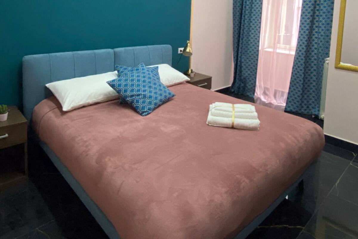 A modern bedroom with a large bed, teal walls, and a window with patterned curtains.