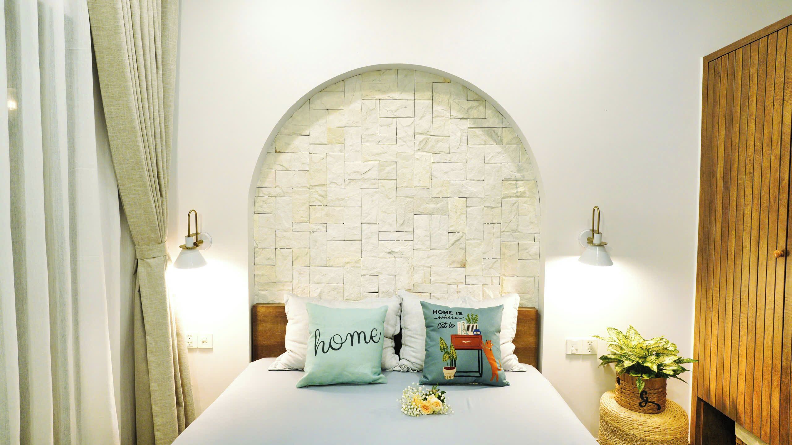 A bedroom with a stone headboard, two bedside lamps, a bed with pillows, a small plant, and a wooden wardrobe.