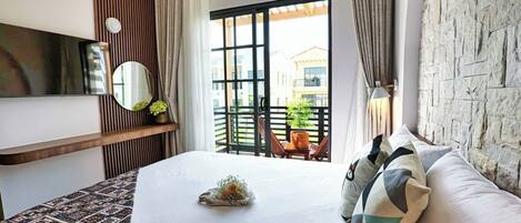 A modern hotel room with a large bed, a wooden shelf with a mirror and a potted plant, a stone-textured wall, and a balcony with a view of buildings.