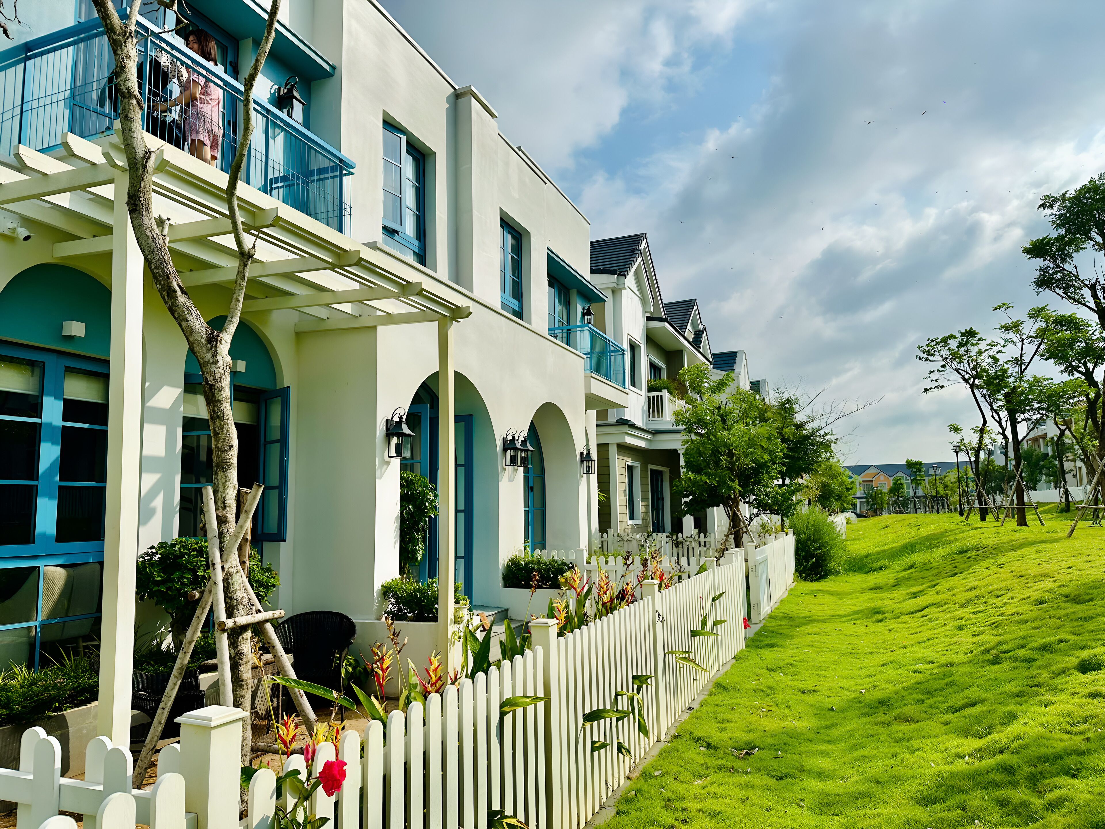A row of modern houses with balconies, white picket fences, and lush green lawns.