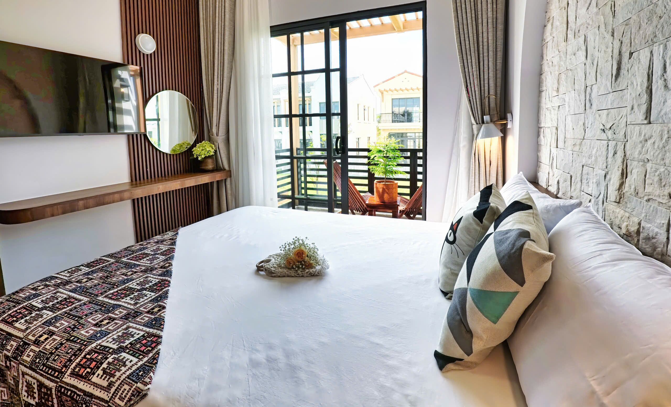 Habitación de hotel con una cama, un estante de madera con un espejo y una planta en maceta, vista al balcón exterior y una pared con textura de piedra.