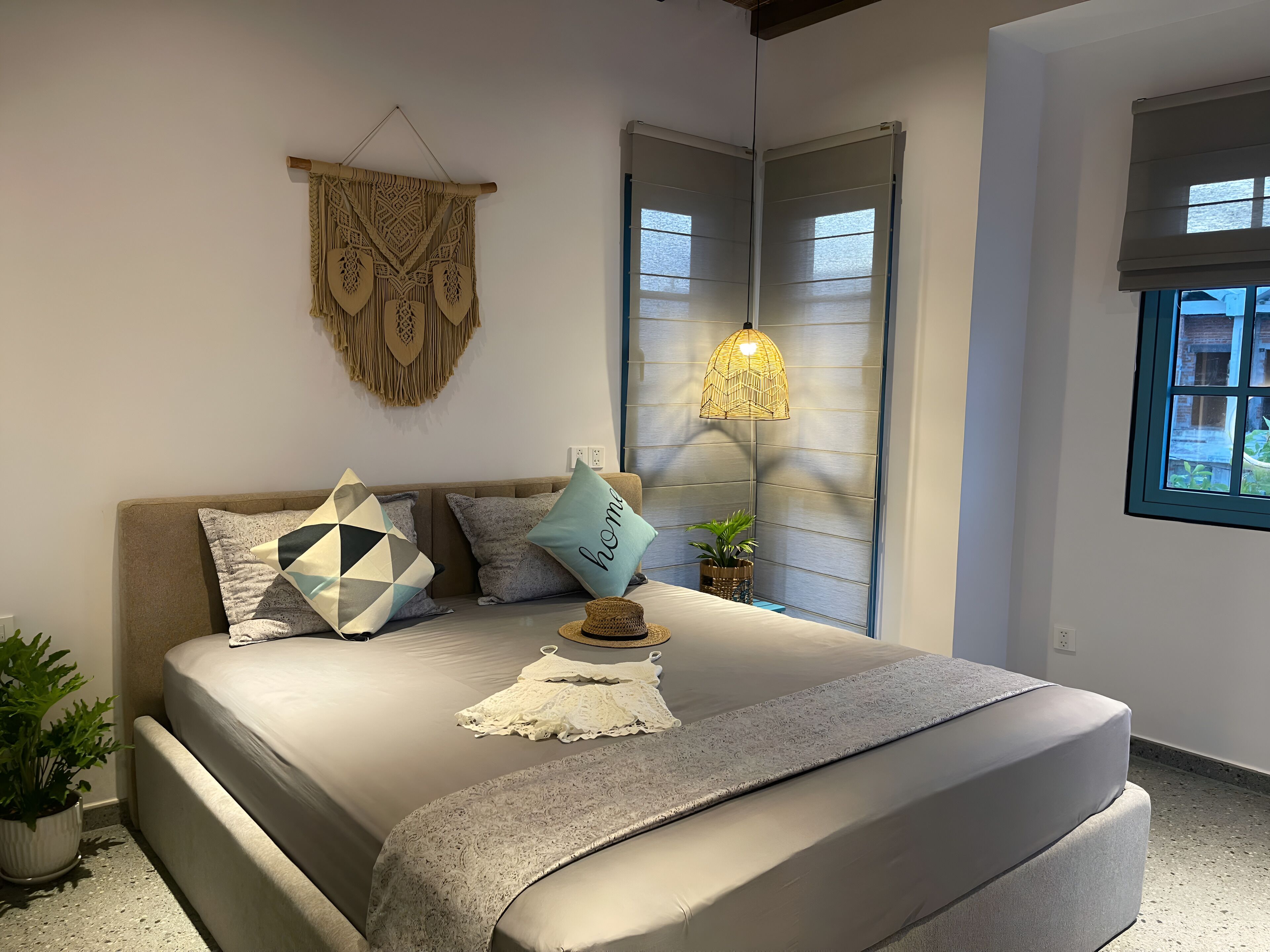 Un dormitorio con cama, almohadas, un tapiz de macramé, una lámpara de pie y una ventana con persianas.