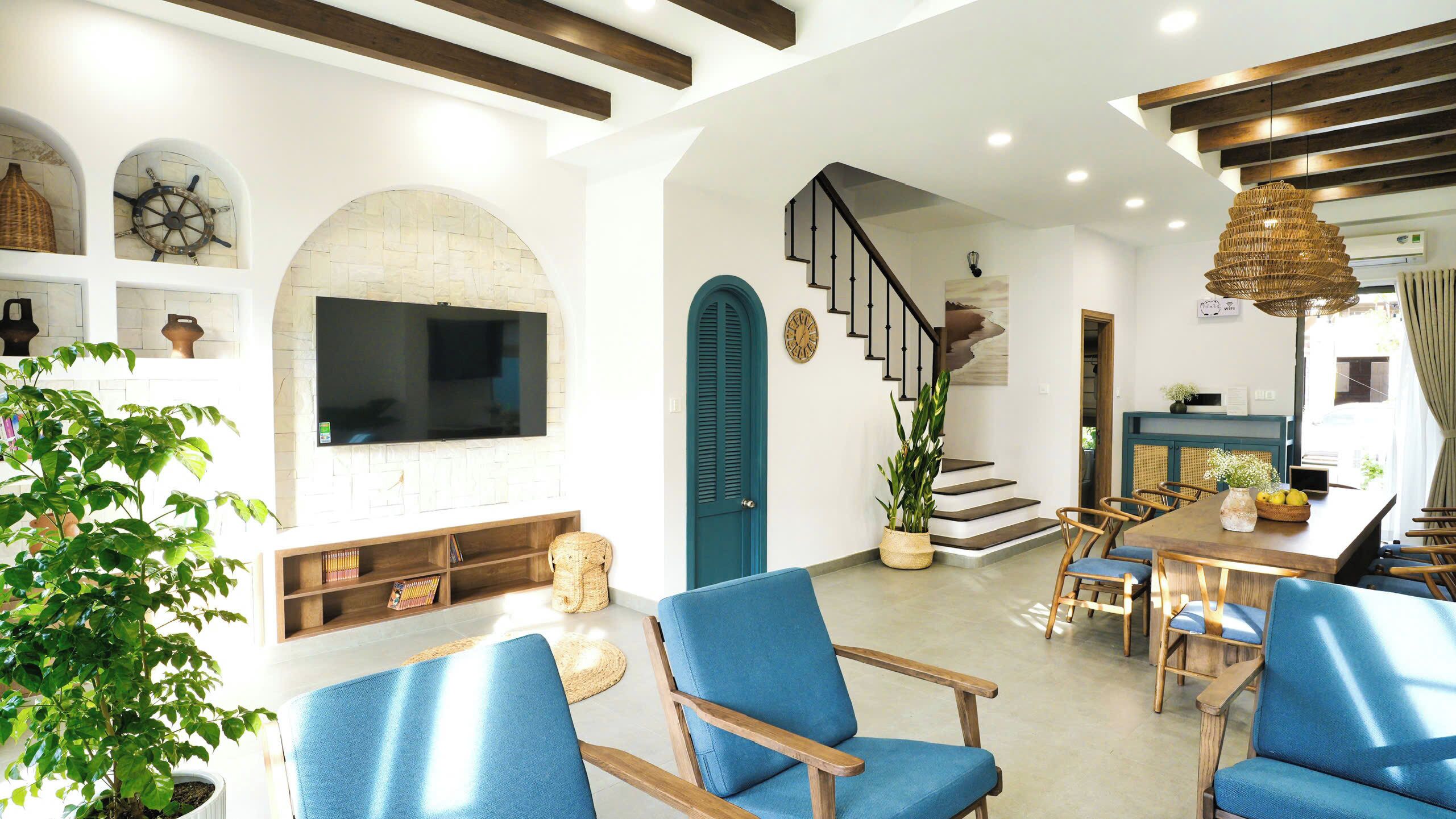 A modern living room with a large flat-screen TV, blue upholstered chairs, a staircase, and a dining area with wooden furniture.