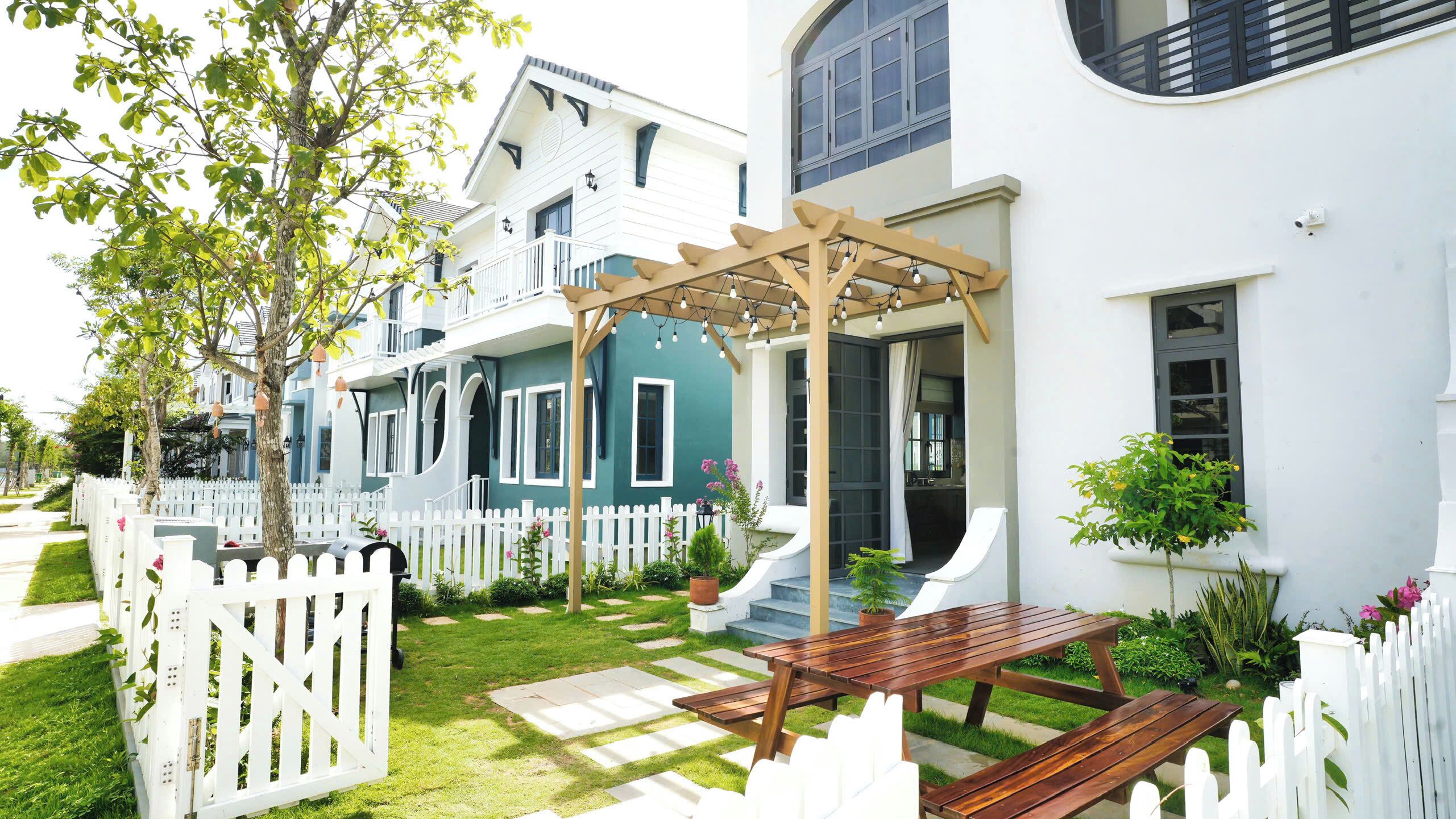 A two-story house with a white picket fence, a wooden pergola with string lights, a wooden bench, and a small garden area.