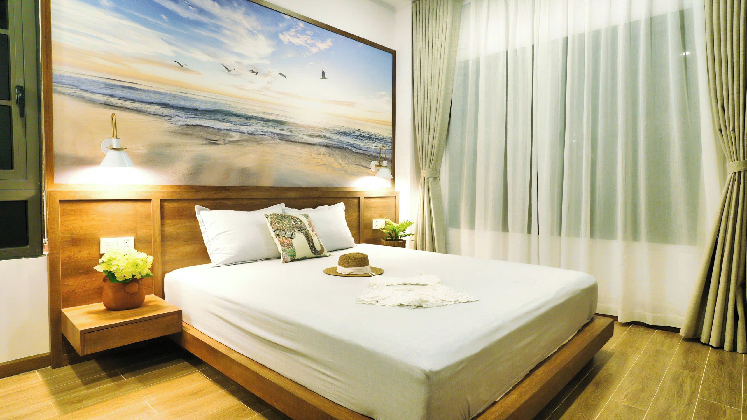 Een hotelkamer met een groot bed, een houten hoofdeinde en een schilderij van een strand. Er staan twee nachtkastjes met lampen en een potplant.