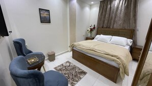 A bedroom with a bed, two chairs, a small table, a nightstand, a bedside table, a mirror, and a window with curtains. - Amadeus familias (Giza)