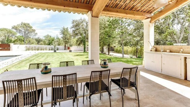 An outdoor patio with a wooden table and chairs, a pergola overhead, and a view of a grassy yard and trees.
