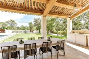 En uteplats med ett träbord och stolar, en pergola ovanför och utsikt över en gräsklädd trädgård och träd.