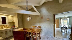 A spacious kitchen with a large island, wooden chairs, and a dining table. There is a clock on the wall and a pendant light above the island. - New " Villa Nova " (Cornillon Confoux)