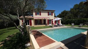A swimming pool with a wooden deck, a house with red shutters, and a patio with lounge chairs. - New " Villa Nova " (Cornillon Confoux)