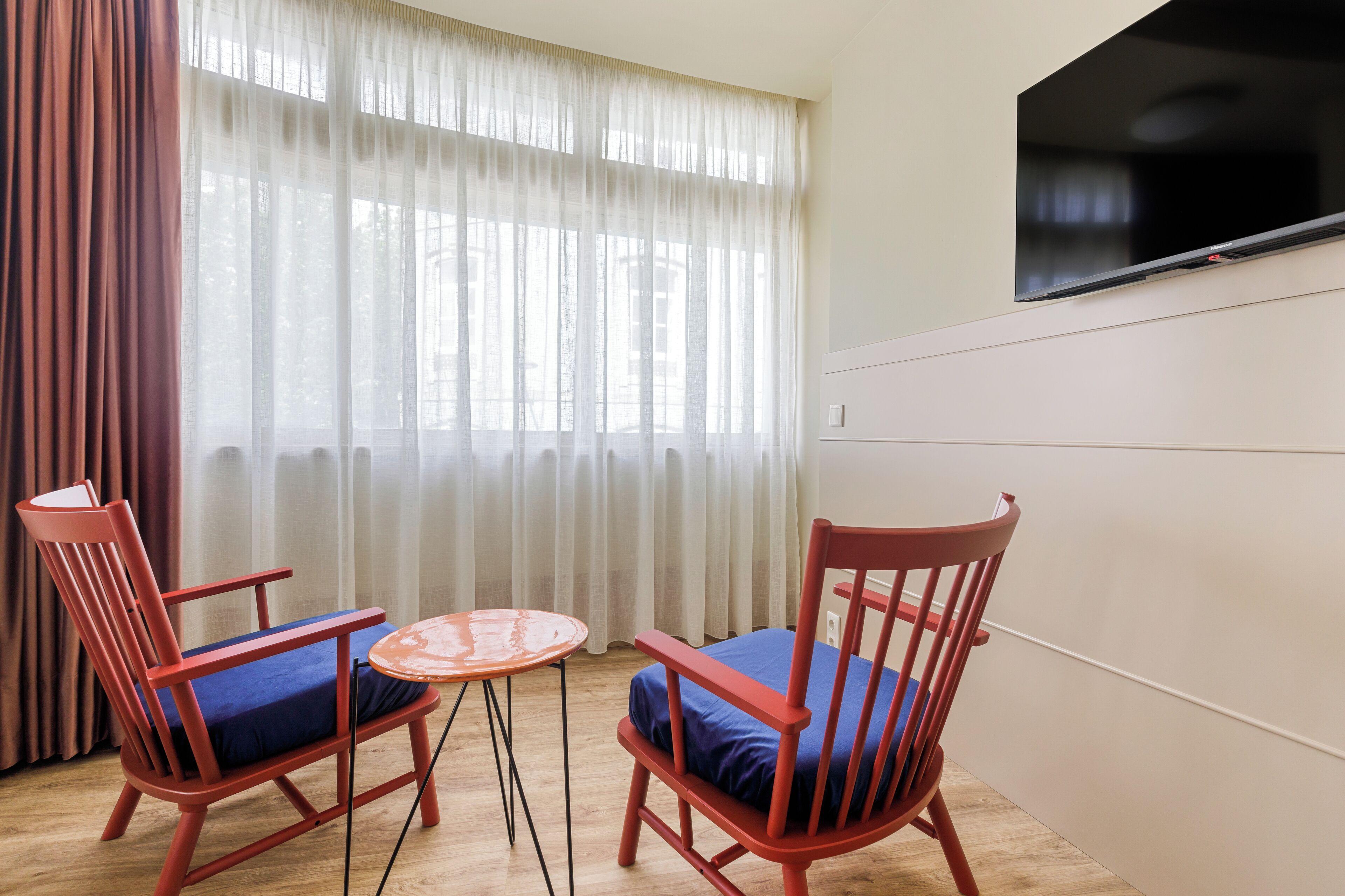 Een kamer met twee rode stoelen, een kleine ronde tafel en een groot raam met dunne gordijnen.