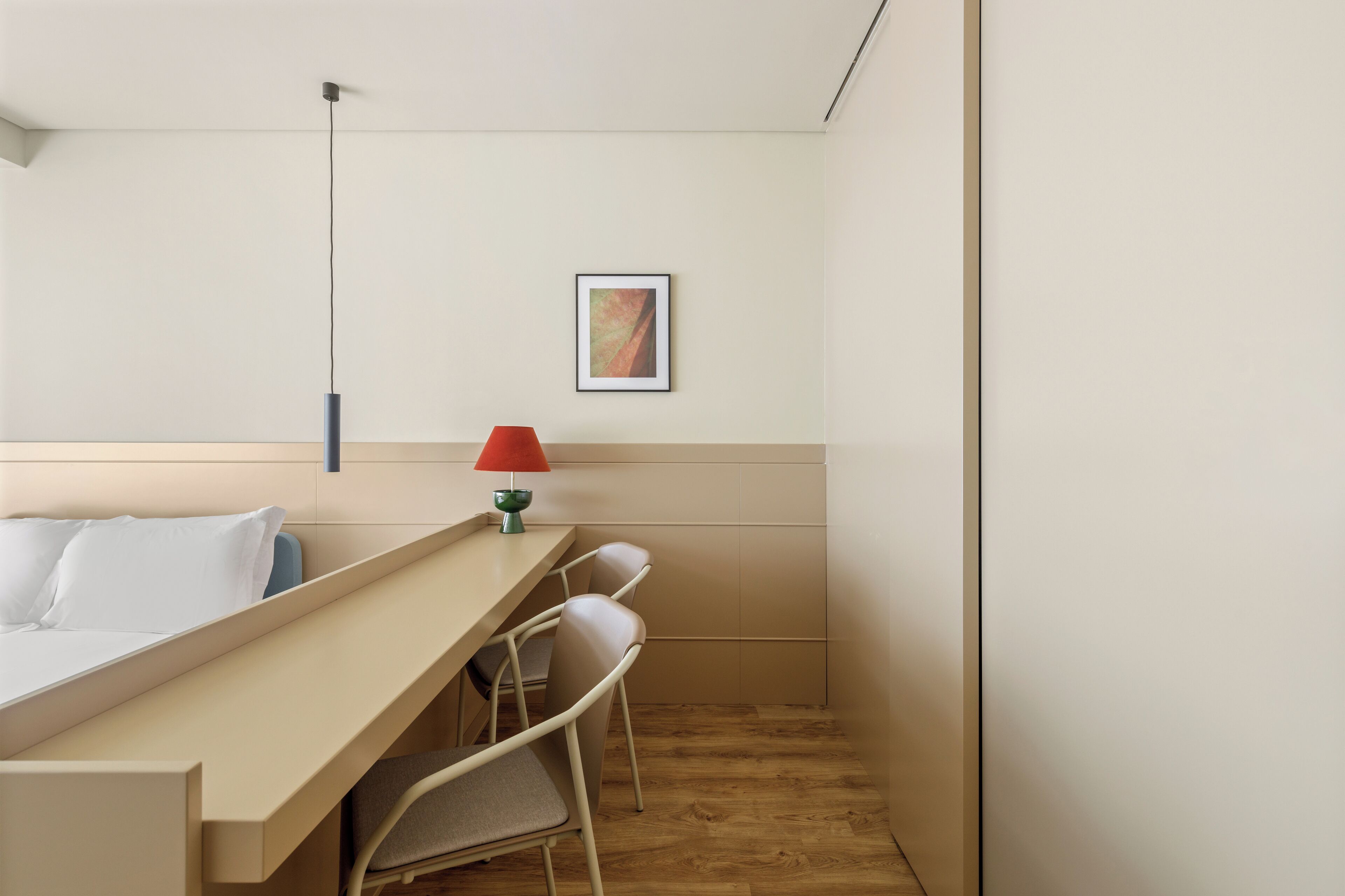 A small, modern hotel room with a bed, a desk, two chairs, a red lamp, and a framed picture on the wall.