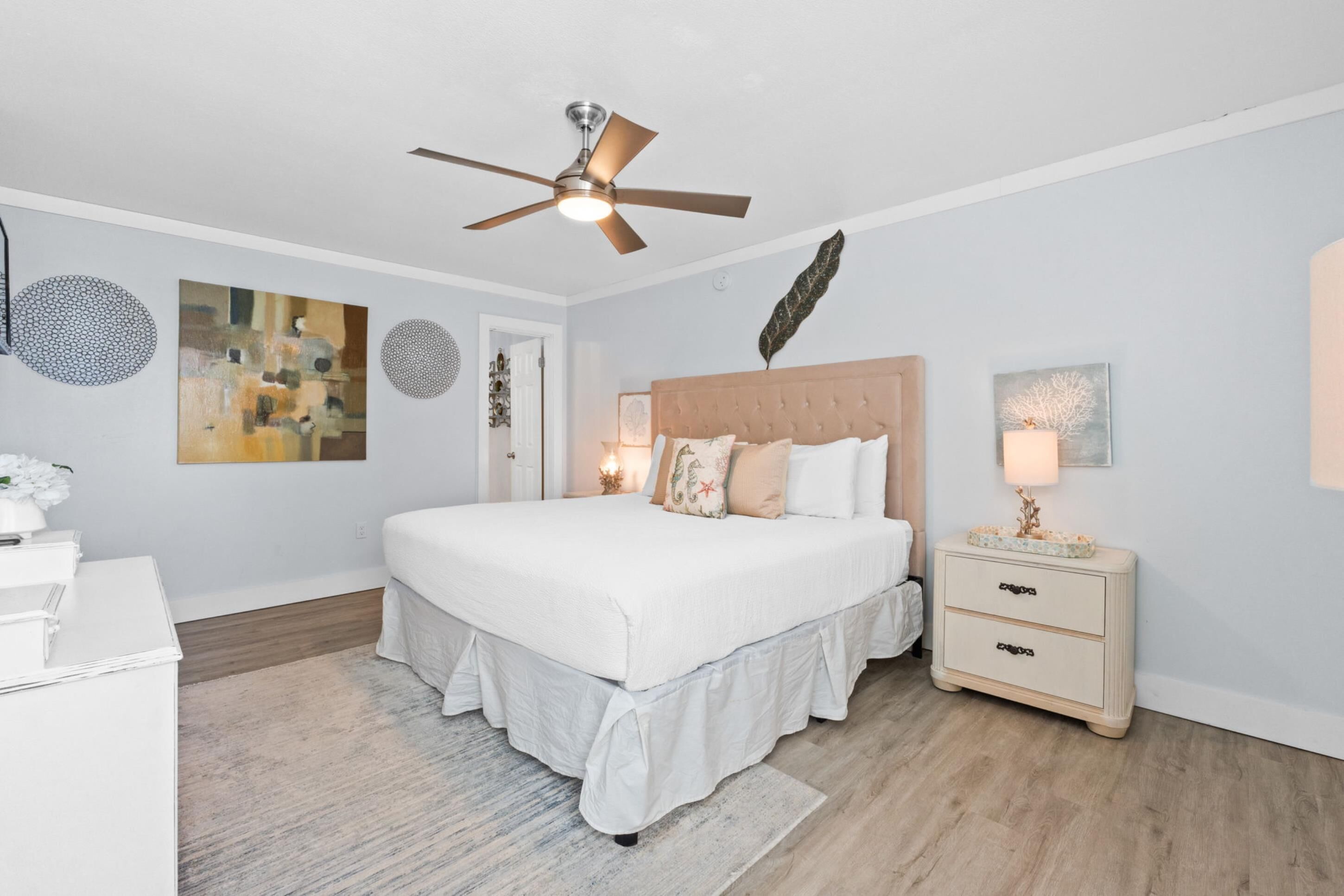 A bedroom with a large bed, a ceiling fan, a nightstand with drawers, and artwork on the walls.