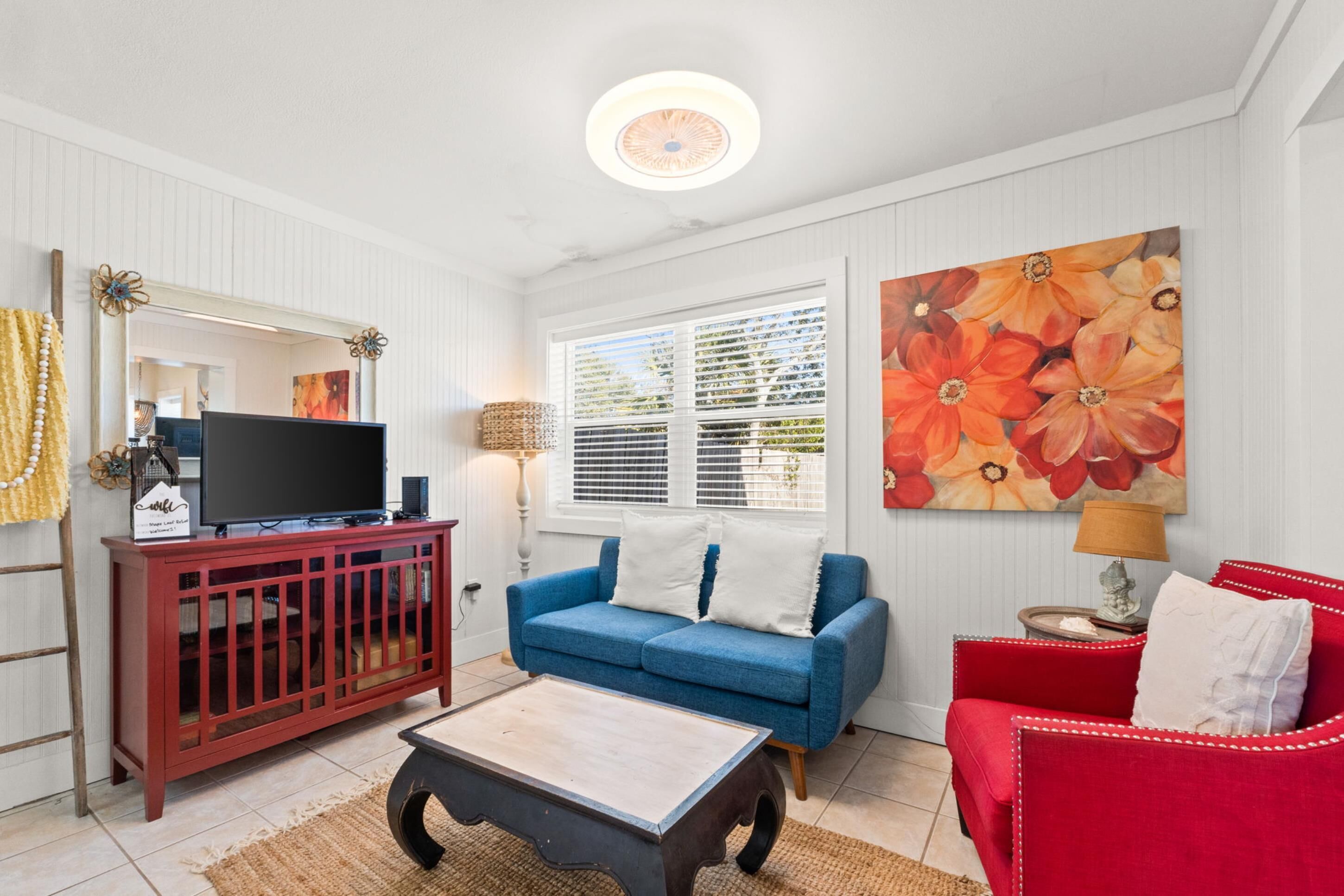 Een woonkamer met een televisie op een rode kast, een blauwe bank, een rode fauteuil en een groot bloemen schilderij aan de muur.