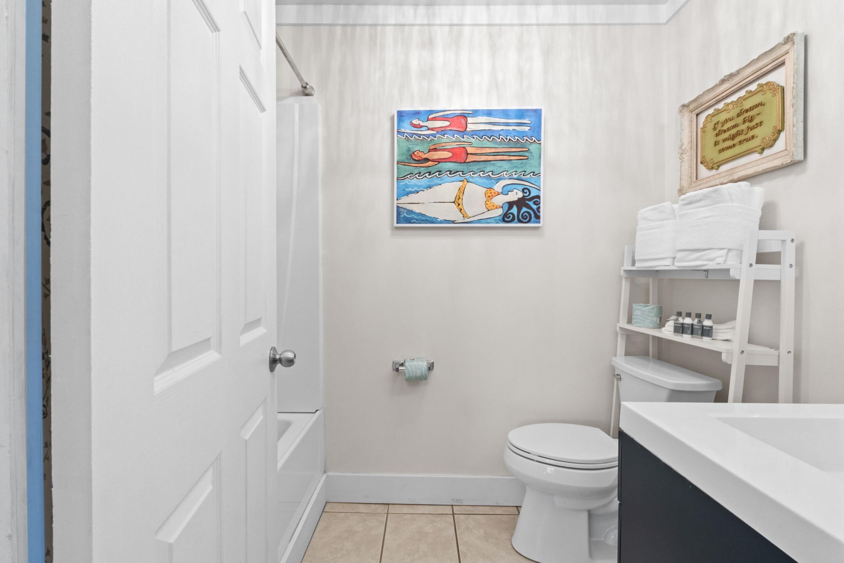 A bathroom with a white toilet, a sink with a white countertop, a towel rack with folded towels, a framed quote on the wall, and a painting of swimmers.