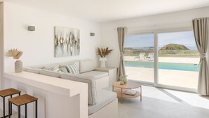 A modern living room with a sofa, a coffee table, and a view of a pool and beach. - Villa Lobelia - Ionian Villa Collection (Lixouri)