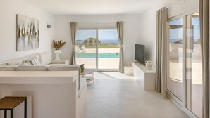 A spacious living room with a large window, a flat-screen TV, and a view of a beach and pool. - Villa Lobelia - Ionian Villa Collection (Lixouri)