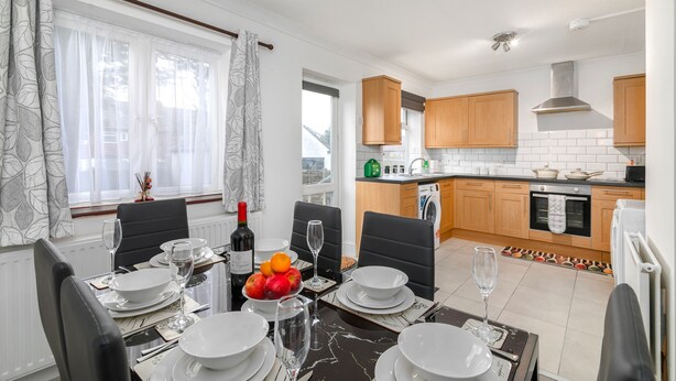 A dining area with a table set for four, a kitchen in the background, and a window with curtains. - Refurbished 4-bed Family Home-near Central London (London)