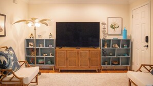 A living room with a television, wicker chairs, a blue shelving unit with decorative items, and a white door. - 2BR/2.5BA in the PERFECT location in Ocean Springs! Downtown Sanctuary (Ocean Springs)