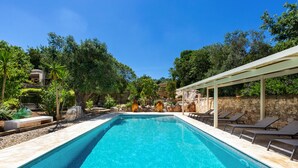 A rectangular swimming pool with clear blue water, surrounded by a stone wall and a pergola with lounge chairs.