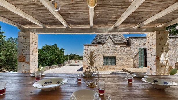 An outdoor dining area with stone pillars, wooden ceiling, and a view of a stone building and trees.