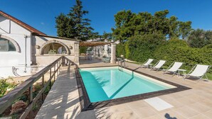 Una piscina con muelle de madera y sillones de descanso, rodeada de vegetación y un edificio con ventanas arqueadas.