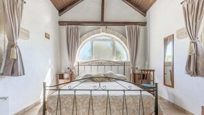Un dormitorio con cama, ventana, cortinas, mesita de noche, espejo y techo de madera.