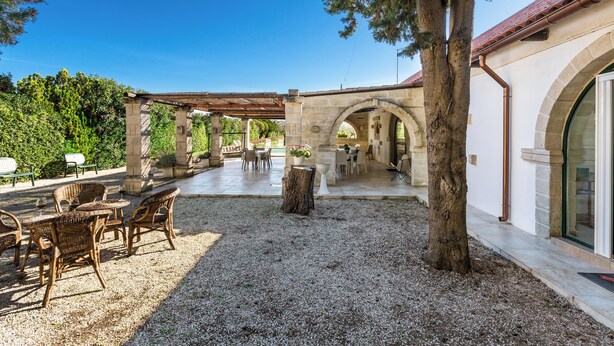 Un patio al aire libre con muebles de mimbre, una pérgola y un arco de piedra que conduce a un espacio de comedor cubierto.