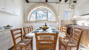 Un comedor con una mesa blanca, seis sillas de madera, un ventanal con vistas al cielo y una cocina con armarios blancos y una estufa.